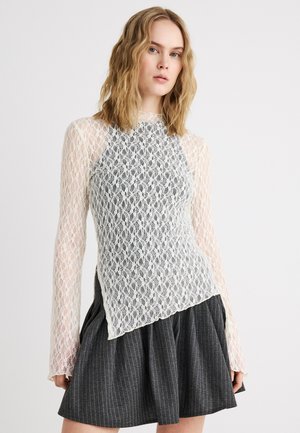 Νεαρή γυναίκα με κυματιστά ξανθά μαλλιά, φορώντας λευκό δαντελένιο μακρυμάνικο τοπ πάνω από μαύρη φούστα με ρίγες, μπροστά σε απλό φωτεινό φόντο.