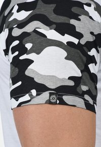 Kurzarmhemd mit einem schwarz-grau-weißen Camouflage-Muster. Verfügt über einen runden Ausschnitt und ein Etikett am unteren Saum. Material aus Baumwollmischung.