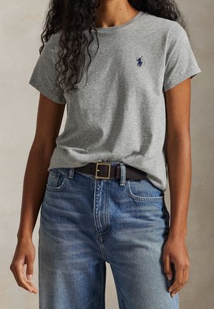Vrouw draagt een grijs T-shirt met korte mouwen en een klein logo, en blauwe hooggesneden spijkerbroek met een zwarte riem.
