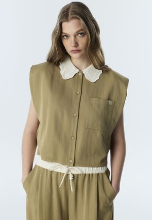 Jeune femme aux longs cheveux ondulés portant un haut olive sans manches boutonné avec col blanc festonné et pantalon assorti, mains dans les poches.