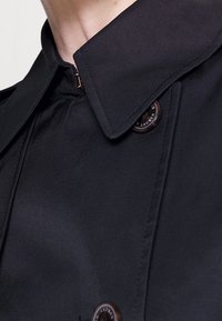 Svart trenchcoat med hög krage, försedd med framdragkedja och bruna knappar. Slät tygtextur med rena linjer och skarpa detaljer.