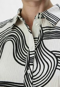Chemise à boutons fabriquée dans un tissu léger, avec des motifs abstraits de vagues noires sur un fond blanc ; comprend un col classique et des boutons.