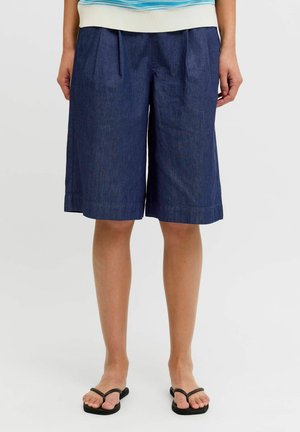 Frau trägt lockere dunkelblaue knielange Shorts und schwarze Flip-Flops, steht vor einem schlichten hellen Hintergrund.