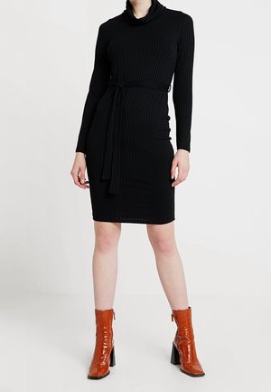 Vrouw draagt een zwarte geribbelde jurk met lange mouwen en een tailleceintuur, gecombineerd met bruine enkellaarsjes met hoge hakken, staand tegen een effen achtergrond.