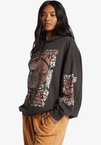 Sudadera negra de gran tamaño con estampado gráfico de un esqueleto, detalles florales coloridos y texto. Fabricada con un tejido suave y un corte relajado, cuenta con un bolsillo canguro.