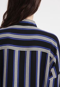 Personne portant une chemise noire avec des rayures verticales et horizontales bleues et blanches, vue de dos avec de longs cheveux foncés visibles.