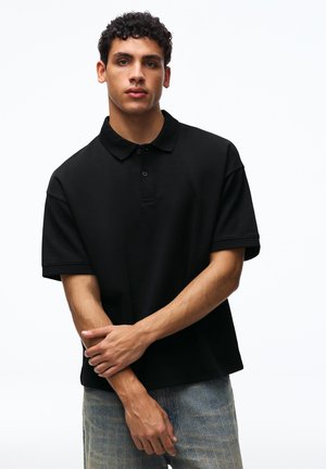 Junger Mann mit lockigem Haar, der ein schwarzes kurzärmeliges Polohemd und hellblaue Jeans trägt, steht mit verschränkten Armen.