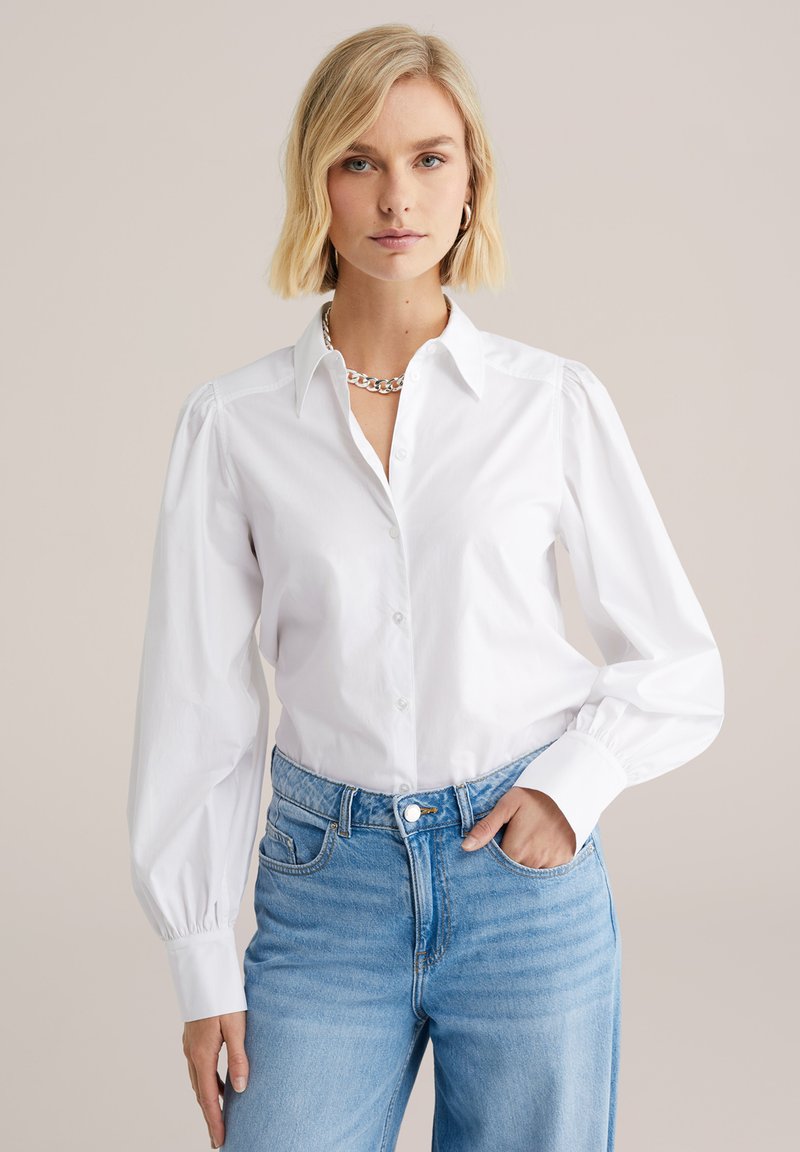 White button-up shirt with long, puffed sleeves, and a pointed collar. Worn with light blue high-waisted jeans. Silver chain necklace accessory.