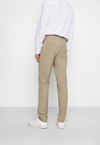 Beige Hosen aus einem glatten Stoff, mit einer taillierten Passform, zwei Gesäßtaschen und getragen mit weißen Sneakers.