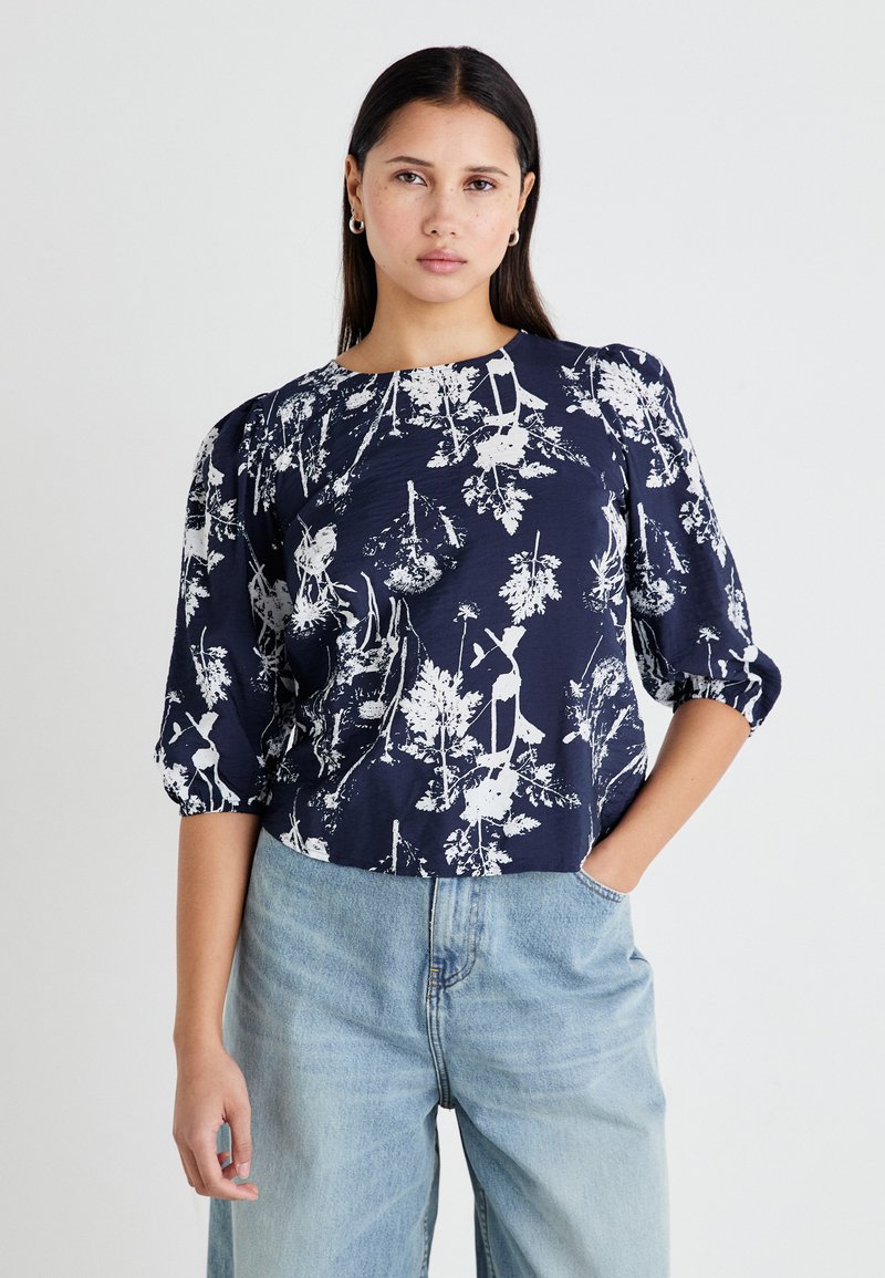 Marinebluse mit weißem botanischem Druck, Rundhalsausschnitt, Puffärmeln und lockerer Passform, kombiniert mit hellblauen weiten Jeans.