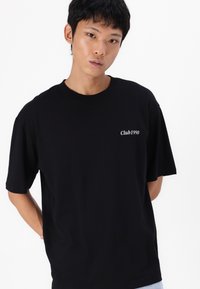 T-shirt en coton noir à manches courtes, avec un petit imprimé blanc "Club1990" sur la poitrine gauche. Design simple et décontracté.