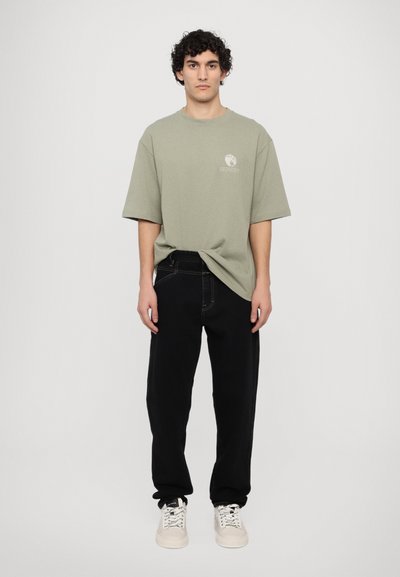 Camiseta oversized verde claro con logo, jeans sueltos negros y zapatillas beige. Tela de algodón con textura suave, sin patrones visibles.
