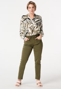 Femme se tenant avec les mains dans les poches, portant un pantalon vert, un chemisier à motifs avec un design de feuilles, des chaussures à brides tressées et des bijoux en or.