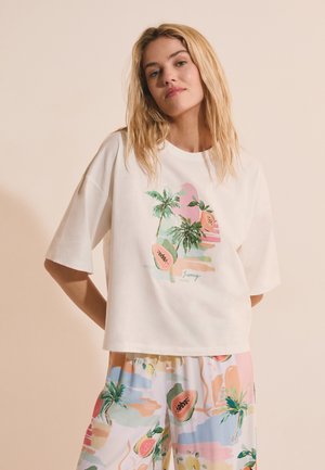 Giovane donna che indossa una t-shirt bianca oversize e pantaloni larghi con stampe colorate di frutta tropicale e palme.