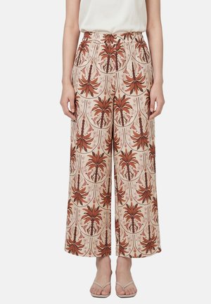 Frau, die hochtaillierte, weit geschnittene Hosen mit braun-beigem Blumenmuster trägt, kombiniert mit weißem Oberteil und beigen Sandalen.