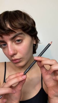 Czarna konturówka do ust MAC trzymana między palcami, lekko zaostrzony koniec w jasnym kolorze mauve. Modelka z krótkimi włosami ubrana w czarny top na ramiączkach.