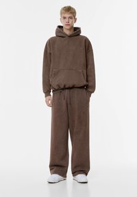 Hasselbruine oversized hoodie met een voorzak en een trekkoord, gecombineerd met bijpassende wijdvallende sweatpants. Witte sneakers maken de outfit compleet.