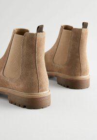 Bottines Chelsea en suede beige avec une surface lisse, des panneaux élastiques sur les côtés et une semelle en caoutchouc robuste avec un léger talon. Anse à l'arrière incluse.