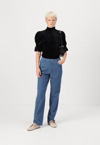 Must musta puff-sleeve top, mis on valmistatud mustast sametist, kombineeritud siniste mustriliste pükstega. Mudel kannab beeži libisevaid kingi ja hoiab väikeses mustas kotis. Lihtne taust.