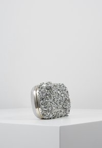 Srebrna kopertówka o teksturowanej powierzchni ozdobionej koralikami i kryształami. O zaokrąglonym kształcie, z metalowym zapięciem i błyszczącymi akcentami.