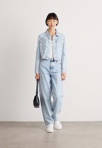 Lyseblå denimjakke med lett slitasje, kombinert med lyseblå høytlivsjeans. Hvit t-skjorte og joggesko, holder en svart veske.