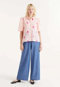 Camisa de manga corta rosa claro con estampado de corazones rosas, combinada con pantalones de mezclilla azul de pierna ancha y bailarina negra. Diseño sencillo de botones en la parte delantera.