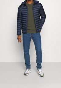 Marineblå puffjakke med quiltet tekstur og hætte, kombineret med en grøn ribstrikket sweater og blå jeans. Hvide sneakers med grå accenter.