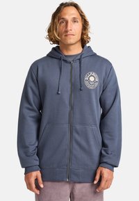 Felpa con zip di colore blu scuro, realizzata in materiale morbido, con un logo sul petto, tasche frontali e lacci regolabili sul cappuccio.