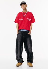 Tricou roșu supradimensionat cu imprimeu alb, blugi negri largi și cizme bej. Purta o șapcă bleumarin și un lănțișor argintiu.