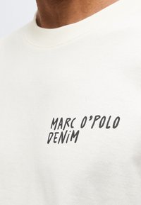 Κοντινό ενός λευκού πουκάμισου με μαύρο κείμενο "MARC O'POLO DENIM" τυπωμένο στο στήθος.