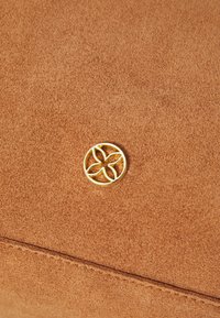 Matériau en daim brun avec un logo circulaire doré arborant un design floral. Texture lisse avec des coutures visibles le long du bord.