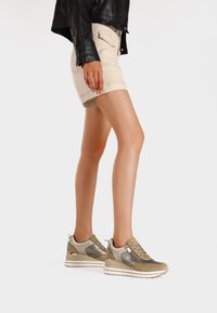 Scarpe da ginnastica beige e verde oliva realizzate con un mix di materiali lisci e testurizzati, caratterizzate da una suola spessa e dettagli con zip, indossate con pantaloni corti beige.