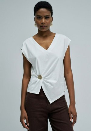 Femme portant un haut blanc sans manches à col en V, torsadé et fixé avec un bouton circulaire, associé à un pantalon marron foncé, sur fond uni.
