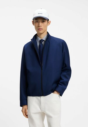 Giovane uomo che indossa un cappellino bianco, giacca blu navy con zip sopra una camicia a righe con cravatta e pantaloni bianchi, in piedi con una mano in tasca.
