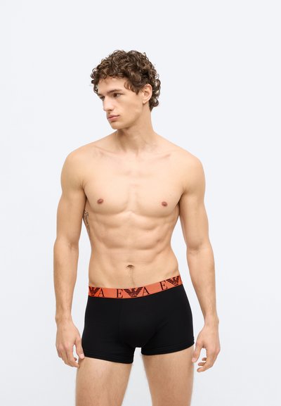 Biancheria intima tipo boxer nera con una fascia in vita con il logo arancione. Il tessuto appare liscio, aderente e si adatta alla forma del corpo di chi lo indossa.