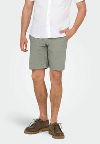 Shorts en coton gris clair pour homme avec une coupe ajustée, associés à une chemise à boutons en lin blanche et des chaussures à lacets en daim marron.