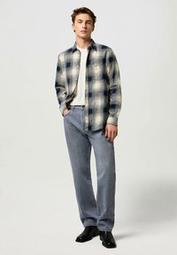 Cămașă din flanel în carouri crem și bleumarin, desfăcută peste un tricou alb, asortată cu blugi bleu deschis cu croială dreaptă și cizme negre.