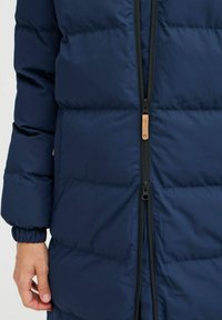 Mornarska puffer jakna z vertikalnim črnim zadrgo, rebrastim manžetami in svetlo rjavim poudarjenjem blizu zadrge; vidna je kviltirana tekstura.