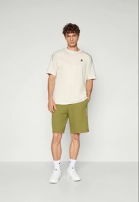 En ung mand står iført en beige Champion t-shirt, olivengrønne shorts, hvide Champion sokker og hvide sneakers foran en ensfarvet baggrund.
