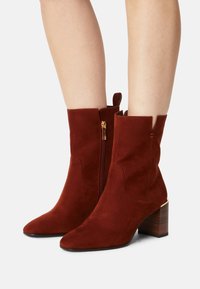 Bottines en daim rouge avec bout pointu, fermeture éclair et talon bloc mi-hauteur orné d'un accent doré.