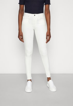 Osoba ubrana w białe, obcisłe, wysokie spodnie typu skinny oraz białe sneakersy, stojąca na tle gładkiego jasnego tła.