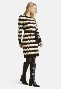 Gebreide jurk met een zigzagpatroon in zwart, room en tan, met een gedefinieerde taille, lange mouwen en een bijpassende riem; gecombineerd met zwarte kniehoge laarzen.