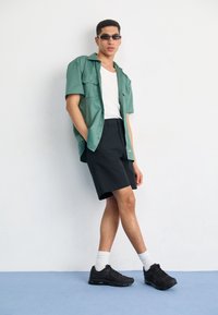 Ung man som bär solglasögon, grön kortärmad skjorta över en vit linne, svarta shorts, vita strumpor och svarta sneakers, stående mot en vit vägg.