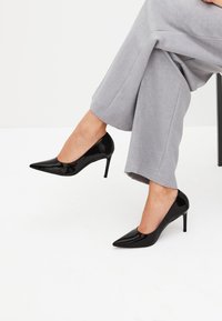 Zwarte lakleren hoge hakken met een spitse neus en een slanke naaldhak, geplaatst naast lichtgrijze, loszittende broeken.