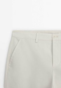 Pantalon beige avec poche latérale, passants pour ceinture, fermeture par bouton et couture nette sur fond blanc.