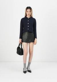 Vrouw in een marineblauwe knoop cardigan, zwarte neplederen minirok, grijze sokken met zwarte puntige hakken, met een zwarte handtas in haar hand.