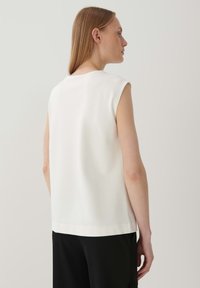 Weißes ärmelloses Oberteil mit runder Halslinie und glatter Textur; getragen mit schwarzen Hosen, was ein sauberes und minimalistisches Design zeigt.