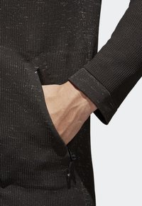 Svart strukturerad långärmad topp med ribbat mönster och blixtlås i sidofickan, med en nära bild av ärmen som visar en åtsittande design.