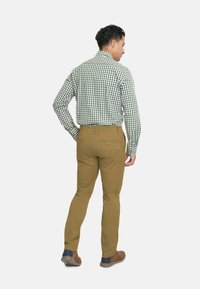 Camisa de manga larga a cuadros verdes y blancos, combinada con pantalones color beige. Bolsillos traseros visibles y zapatos marrones con acentos azules.