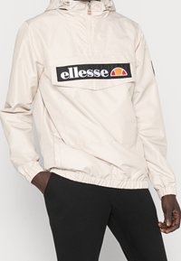 Beige lättviktsjacka med halvdragkedja, elastiska ärmslut och en framficka. Funkar med en svart logotypapplikation med "ellesse."